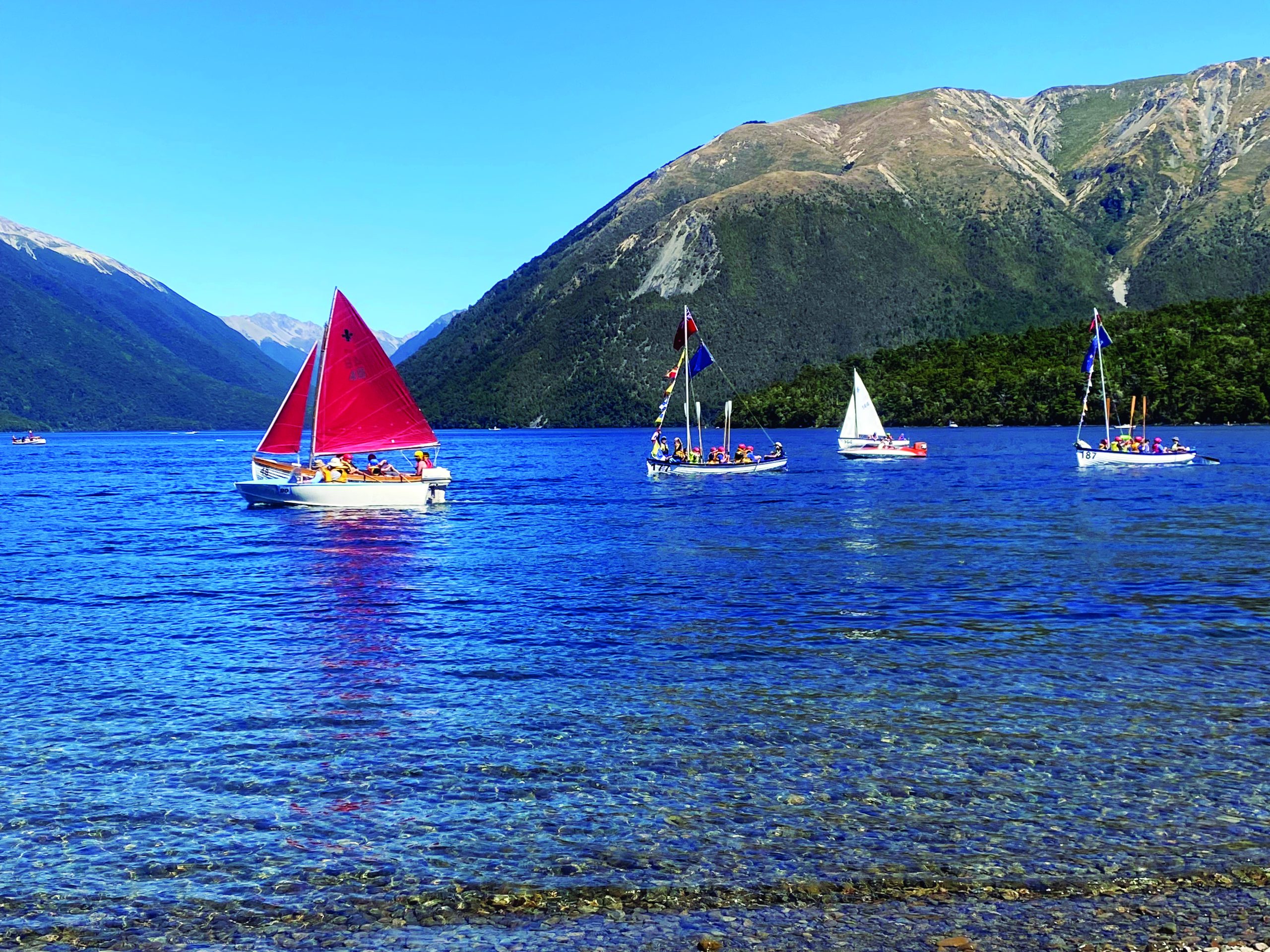 Several vintage sailboats with colorful sails navigate across a pristine lake surrounded by mountains. // Photo credit: supplied.