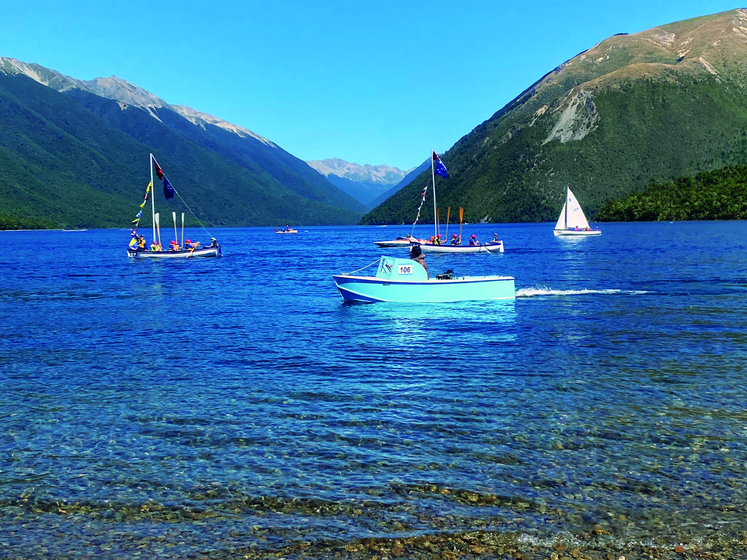 Several boats including a white vessel numbered 106 navigate the crystal-clear blue waters of a mountain-surrounded lake with snow-capped peaks in the background // Photo credit: supplied.