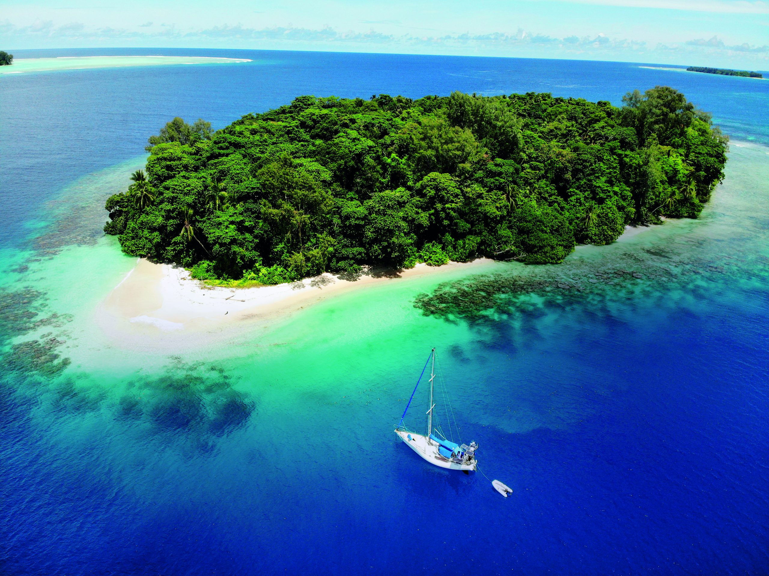 A sailboat anchors in crystal-clear turquoise waters near a pristine tropical island with dense green vegetation and white sandy beaches // Photo credit: BIRGIT HAKL.