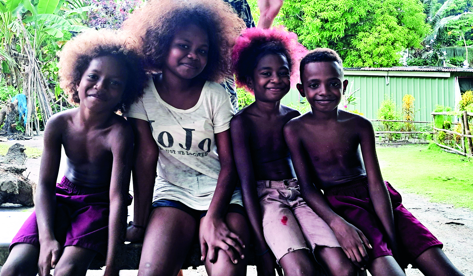 I don't see any boats or boating activity in this image. The photo shows four children sitting together outdoors in what appears to be a tropical or rural setting with lush green vegetation and a building in the background // Photo credit: BIRGIT HAKL.