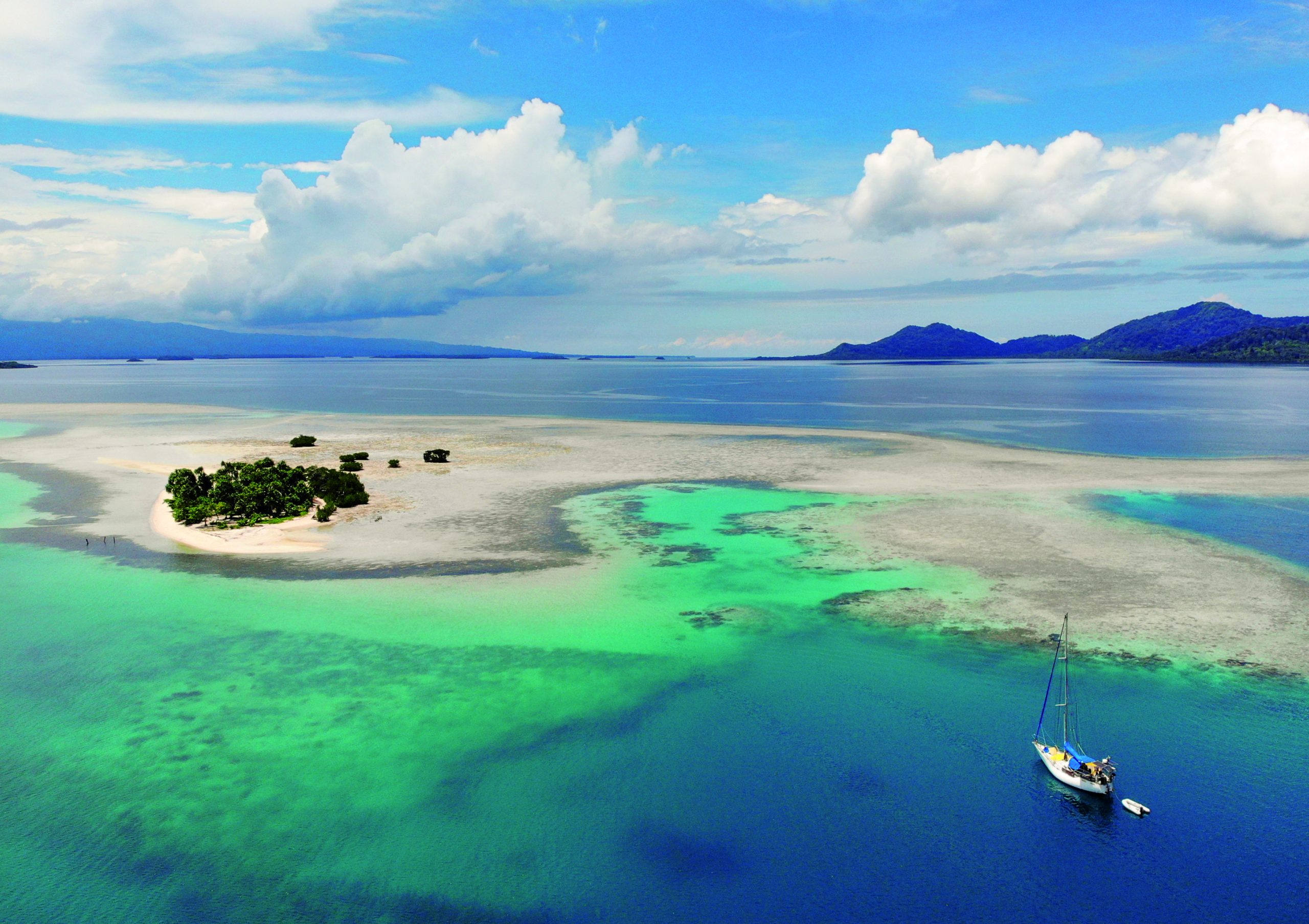 A sailboat anchors in crystal-clear turquoise waters near a small tropical island with white sand beaches and coral reefs visible beneath the surface // Photo credit: BIRGIT HAKL.