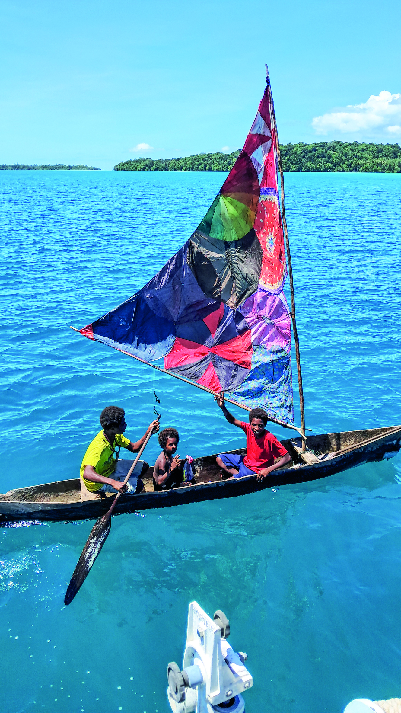 Three people sail a traditional outrigger canoe with a colorful patchwork sail across crystal-clear tropical waters near forested islands // Photo credit: BIRGIT HAKL.