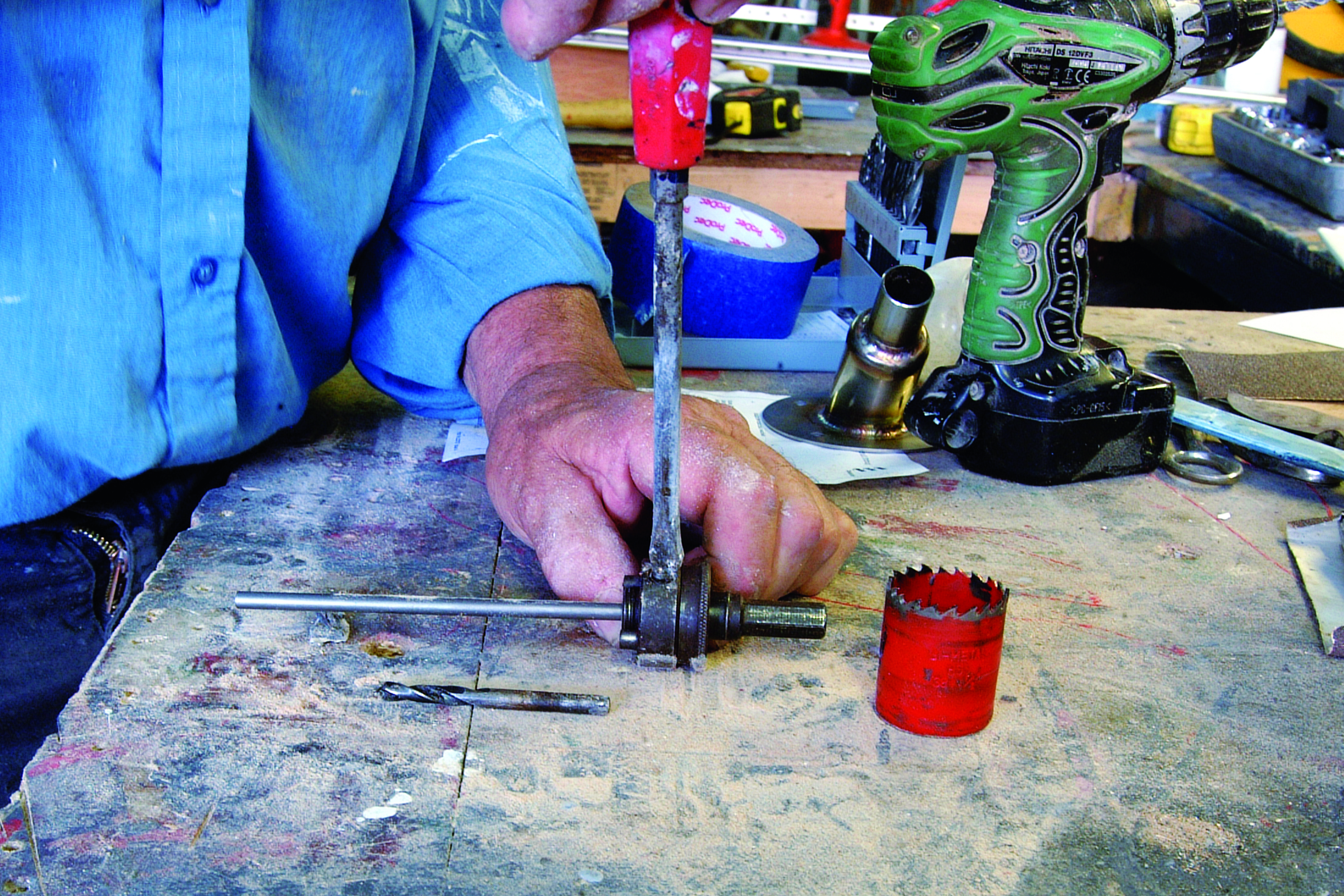 A person uses a hand drill to work on a metal rod at a cluttered workbench with various tools and a red hole saw visible // Photo credit: supplied.