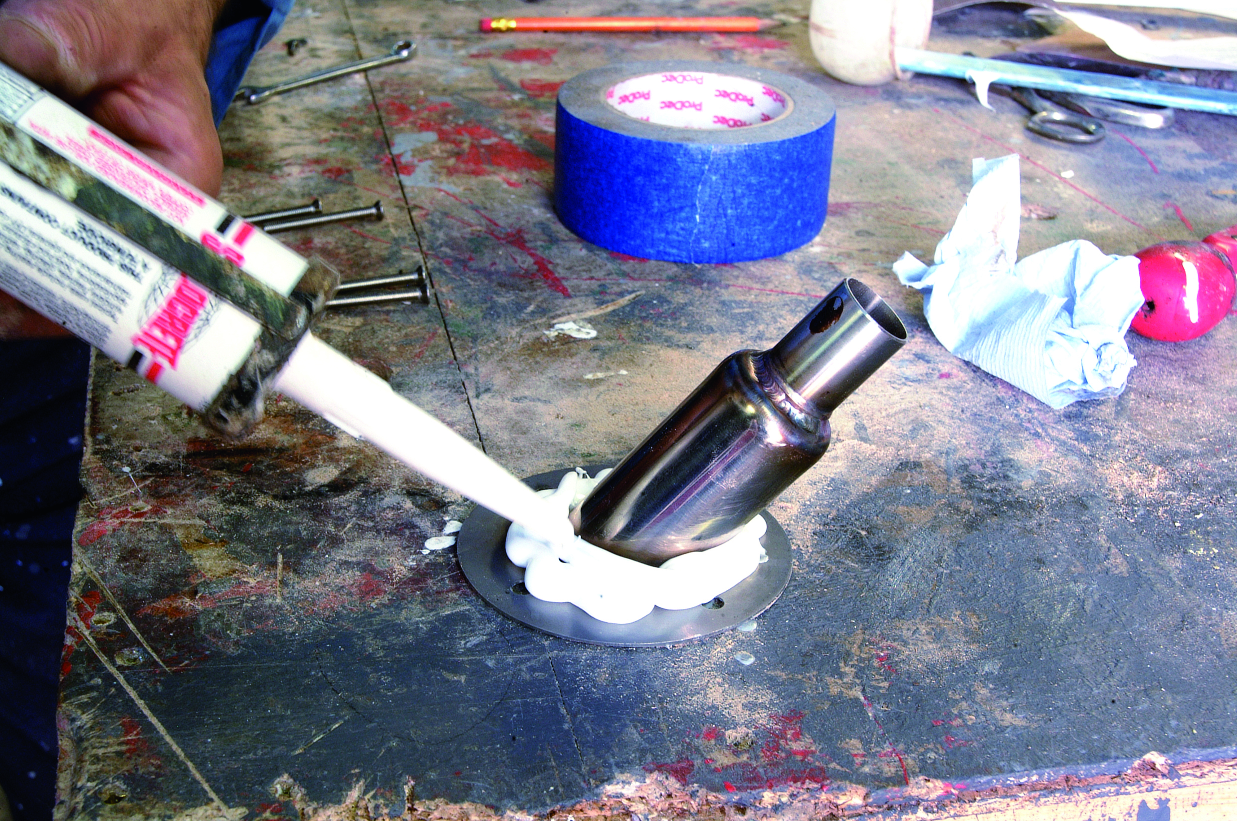 A person applies marine sealant from a caulk gun around a metal through-hull fitting on a boat deck during maintenance work // Photo credit: supplied.