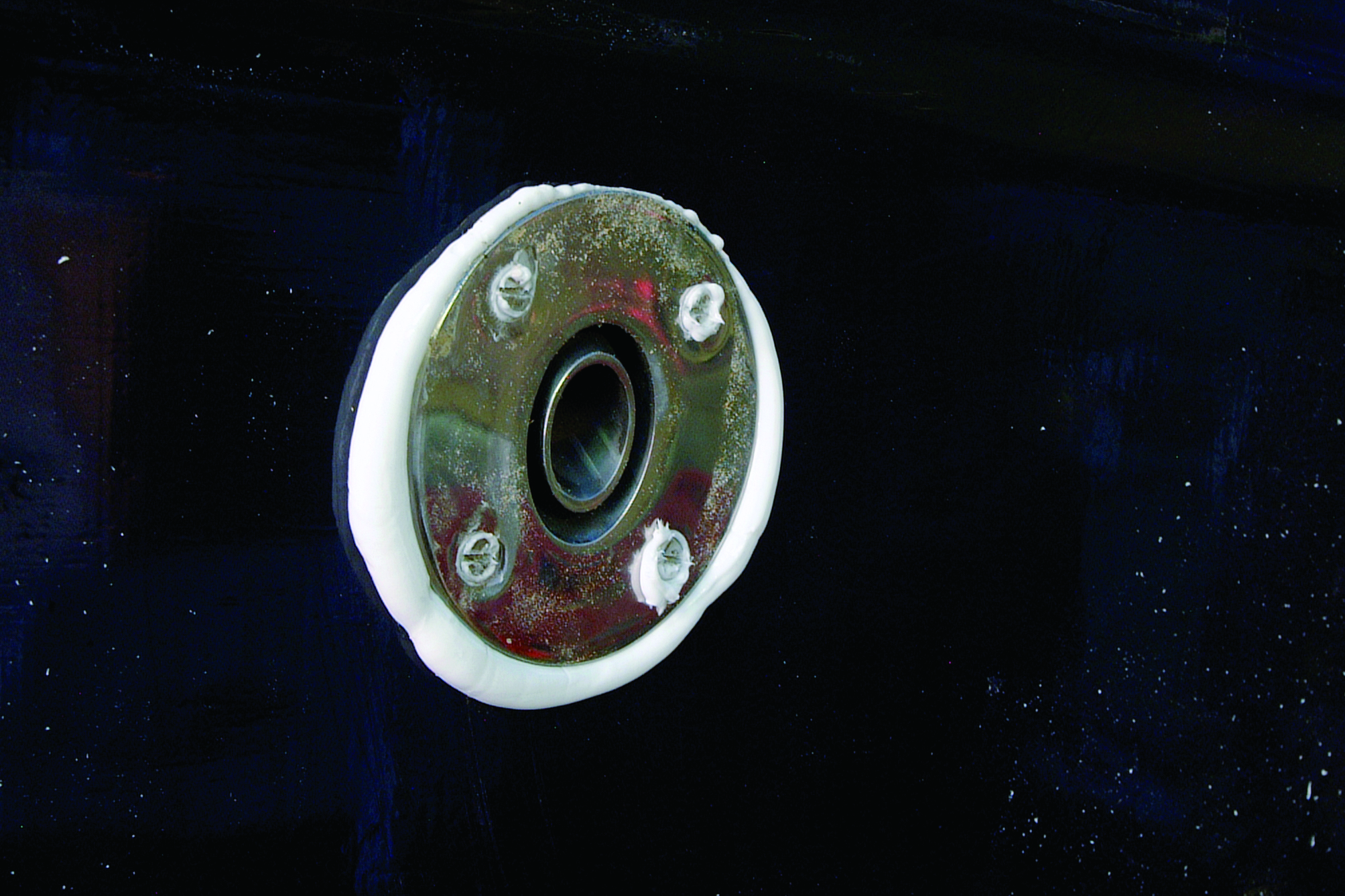 A circular porthole or boat window with a white frame floats in dark blue water, showing reflective glass in the center surrounded by weathered metal with mounting bolts // Photo credit: supplied.
