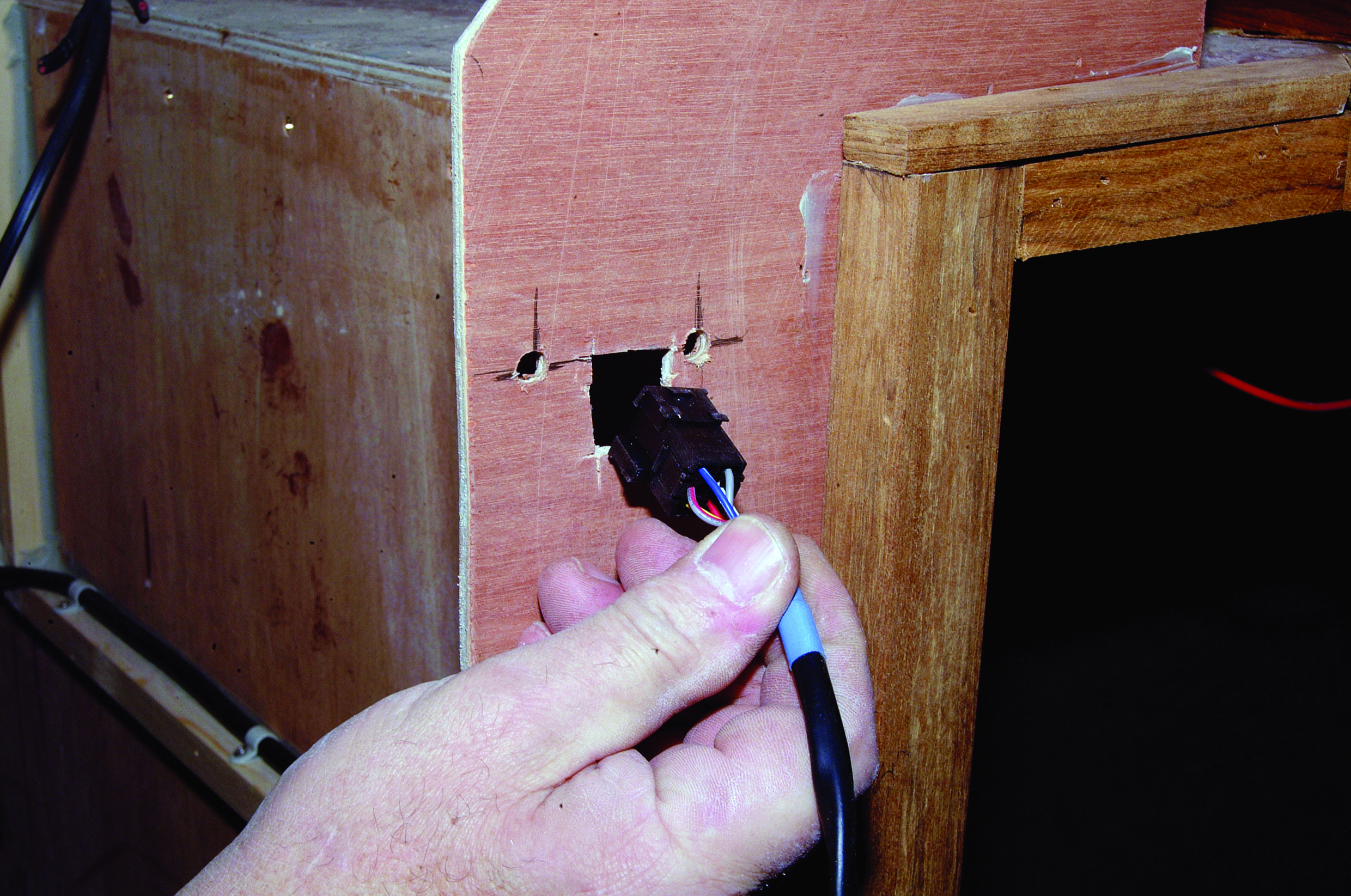 A person's hand holds electrical wires while working on wiring installation through a wooden panel inside a boat cabin // Photo credit: supplied.