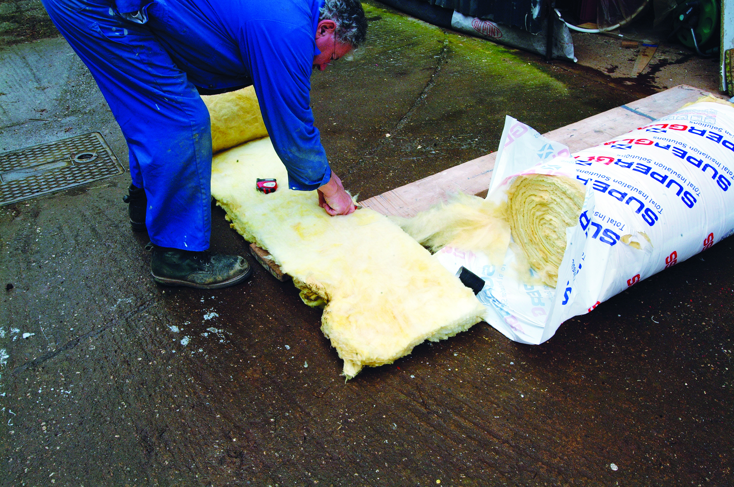 A worker in blue coveralls cuts yellow insulation material from a large roll on a wet dock or marina surface // Photo credit: supplied.