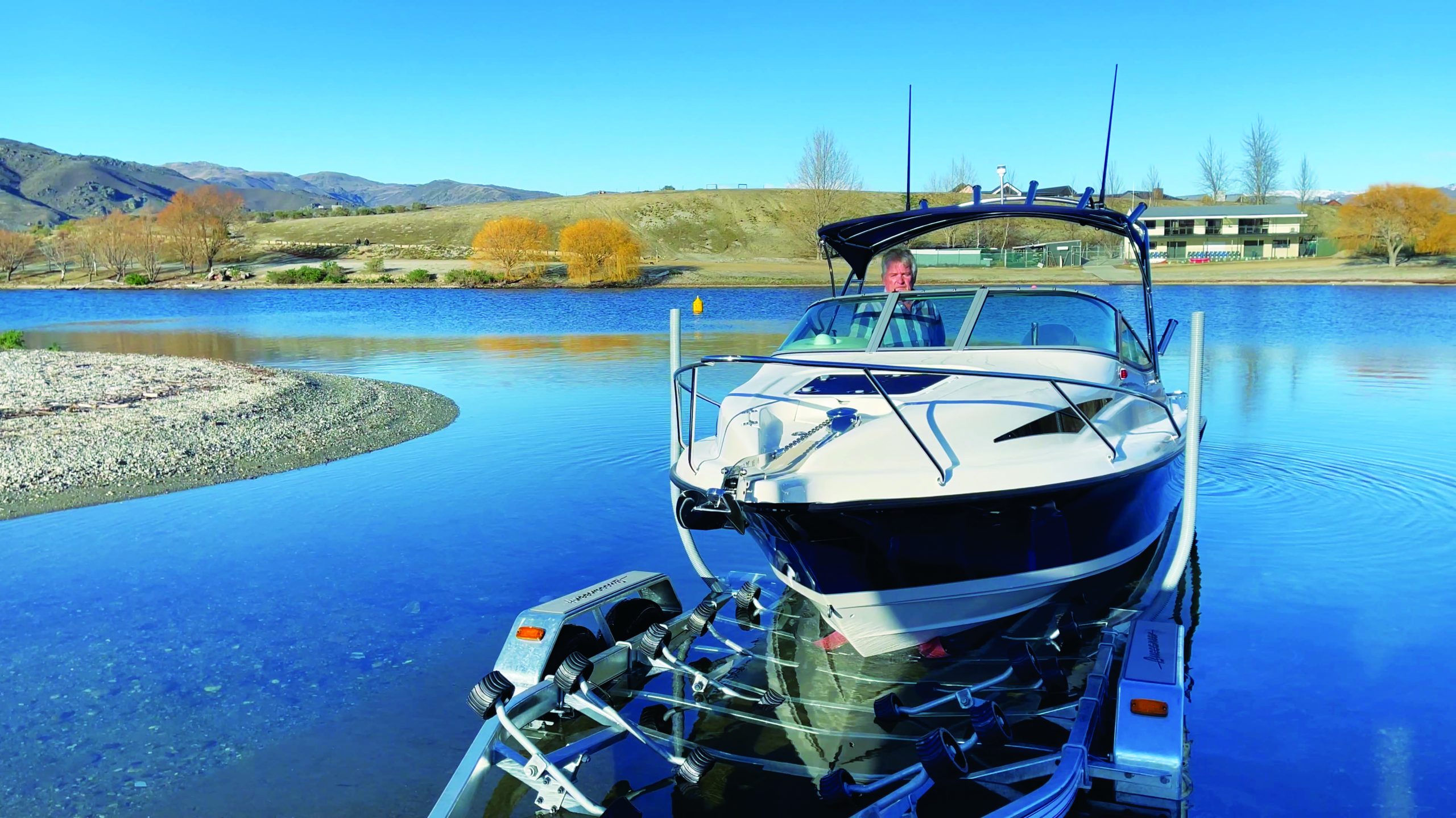 A white and blue powerboat sits on a trailer at a boat launch ramp on a calm lake surrounded by rolling hills and autumn trees // Photo credit: supplied.